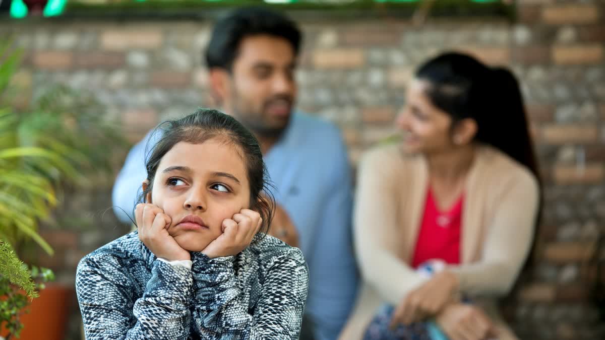 A girl resting her chin in her hands while her parents talk together in the background